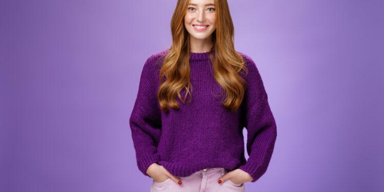 Optimistic attractive ginger girl in 20s wearing purple sweater standing ready and energized over violer background smiling friendly and confident at camera, showing readiness to have fun.