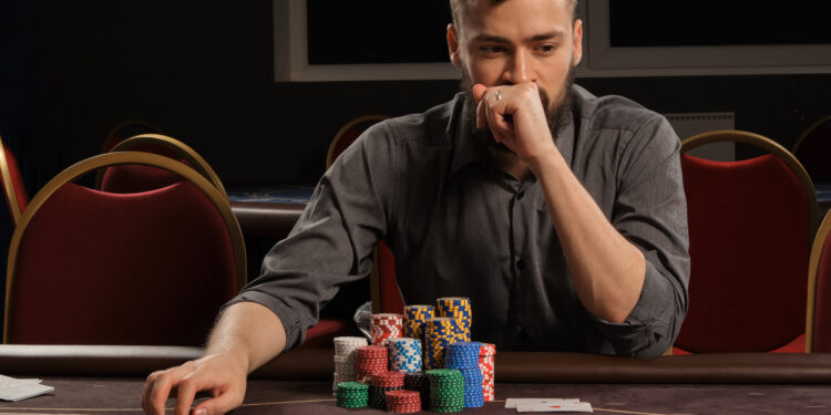 Handsome bearded male in a gray shirt is playing poker sitting at the table in casino. He is making bets waiting for a big win. Gambling for money. Games of fortune.