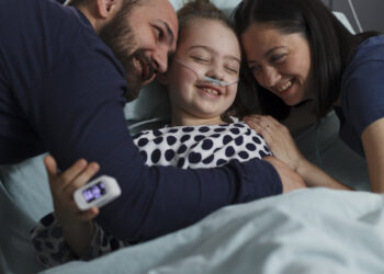 Loving parents hugging sick daughter resting in healthcare clinic pediatrics ward. Happy ill little girl hugged by caring mother and father while sitting with her on patient bed.