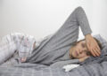 Kanserle Mücadelede Uyku Problemleri 5 Sick young Caucasian woman in grey homewear lying on her side in bed, feeling temperature, touching forehead with hand. Illness, pain concept