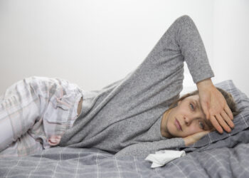 Sick young Caucasian woman in grey homewear lying on her side in bed, feeling temperature,  touching forehead with hand. Illness, pain concept
