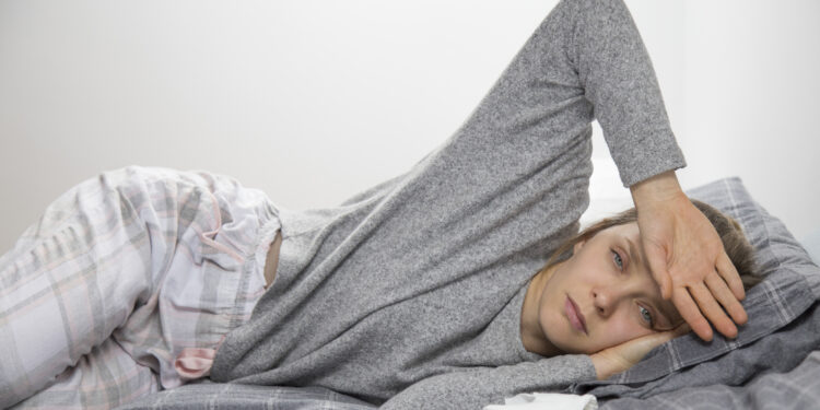 Kanserle Mücadelede Uyku Problemleri 1 Sick young Caucasian woman in grey homewear lying on her side in bed, feeling temperature, touching forehead with hand. Illness, pain concept