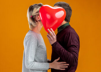 young couple on valentines day covered face with heart balloon isolated on orange background